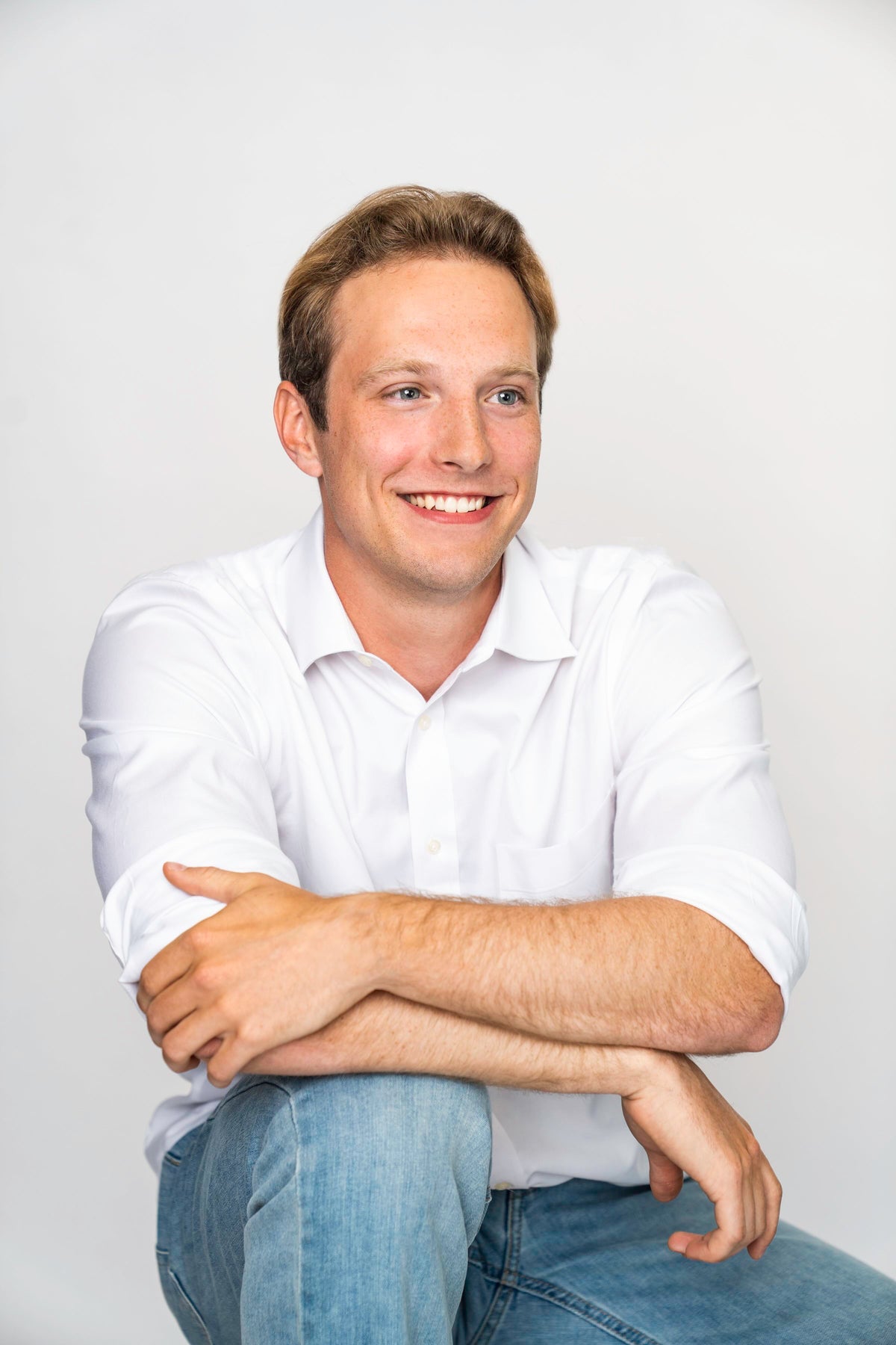 A smiling man with light brown hair wearing a white long-sleeve button-down shirt and blue jeans, sitting with his arms crossed over his knee against a plain light gray background. Alex for Löta Real Food Fiber Co.