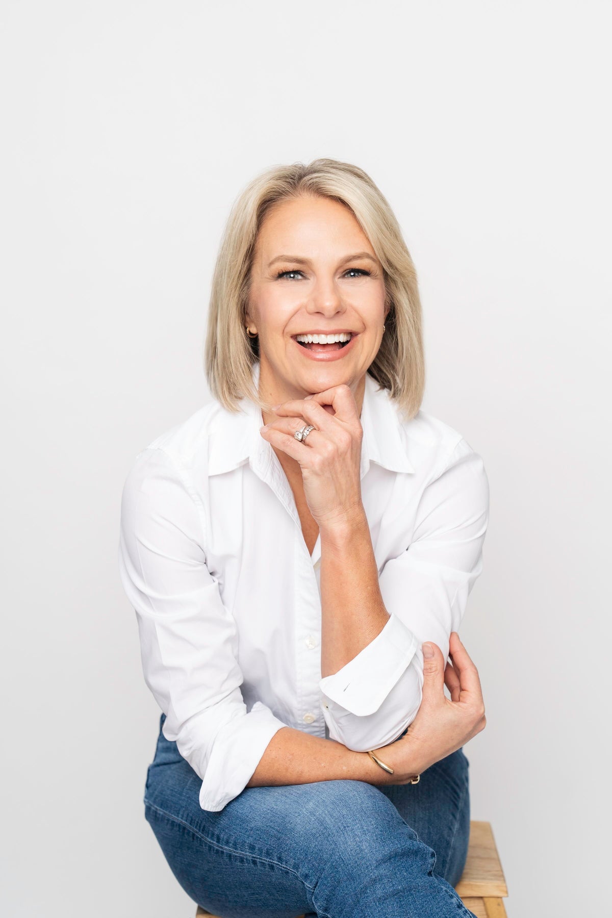 A smiling blonde woman wearing a white button-up shirt and blue jeans sits on a wooden stool against a plain light gray background. Jessica Grelle for Löta Real Food fiber Founder. Nutrition Expert, Food Scientist, Mom
