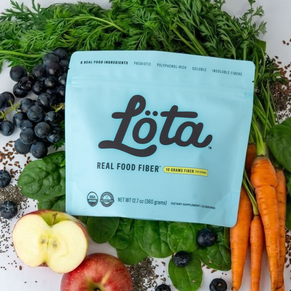 A light blue pouch of Löta Real Food Fiber dietary supplement, surrounded by fresh ingredients including carrots, blueberries, spinach, and sliced apples ORGANIC Löta Real Food Fiber dietary supplement, featuring the brand name 'Löta' in large black script, with 'Real Food Fiber' and '10 Grams Fiber Per Serving' printed below, set on a light surface with a blurred indoor background. Prebiotic Fiber, Polyphenol-Rich Fiber, Soluble Fiber, Insoluble Fiber. Not ultra-processed fiber Lota