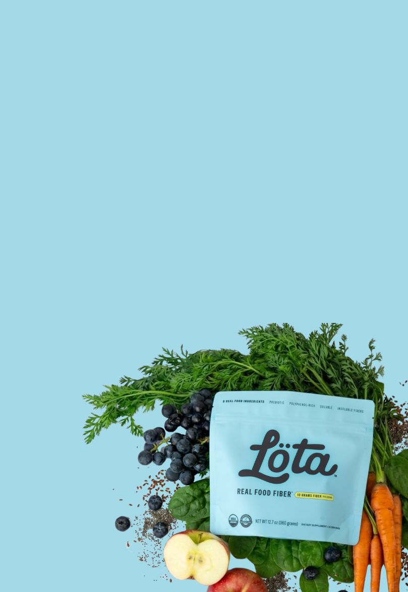 A light blue pouch of Lota Organic Real Food Fiber placed on top of fresh blueberries, spinach, sliced apple, and carrots against a solid light blue background. Lota (Löta)