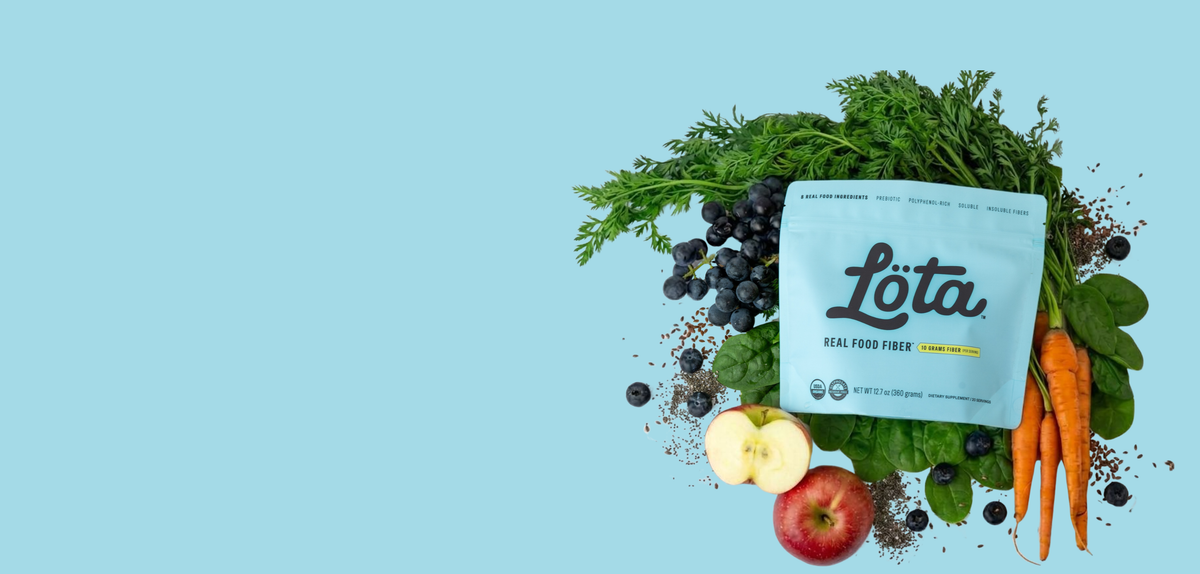A light blue pouch of Lota Organic Real Food Fiber placed on a light blue background, surrounded by fresh fruits and vegetables including an apple, blueberries, spinach, carrots. Not ultra-processed fiber. Lota (Löta)