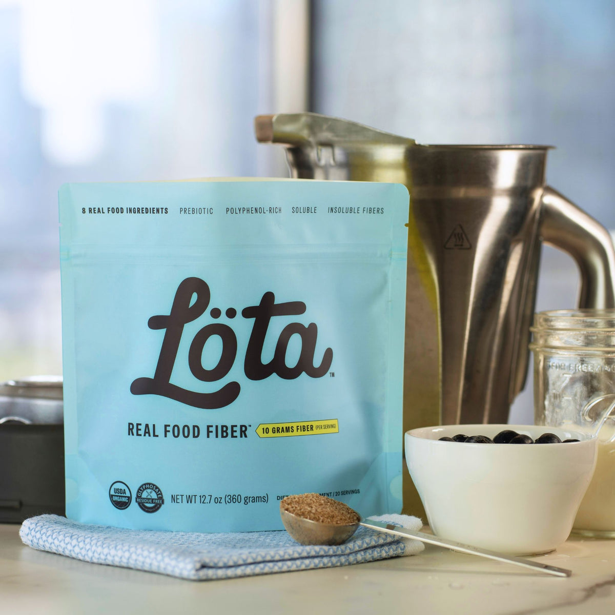 A light blue stand-up pouch of Löta Real Food Fiber granola mix sits on a countertop with a metal blender pitcher, a small bowl of dark berries, and a spoonful of the granola mix in front of it. A light blue stand-up pouch of ORGANIC Löta Real Food Fiber dietary supplement, featuring the brand name 'Löta' in large black script, with 'Real Food Fiber' and '10 Grams Fiber Per Serving' printed below, set on a light surface with a blurred indoor background. Prebiotic Fiber, Polyphenol-Rich Fiber, Soluble Fiber,