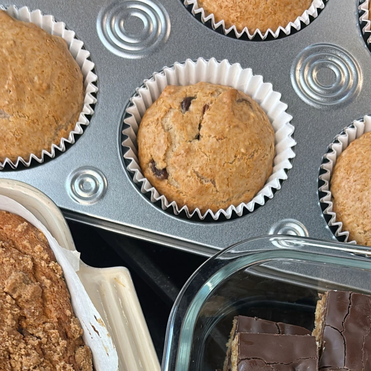 A close-up overhead view of freshly baked chocolate chip muffins in white paper cups inside a gray metal muffin tin, with other baked goods partially visible in the frame.