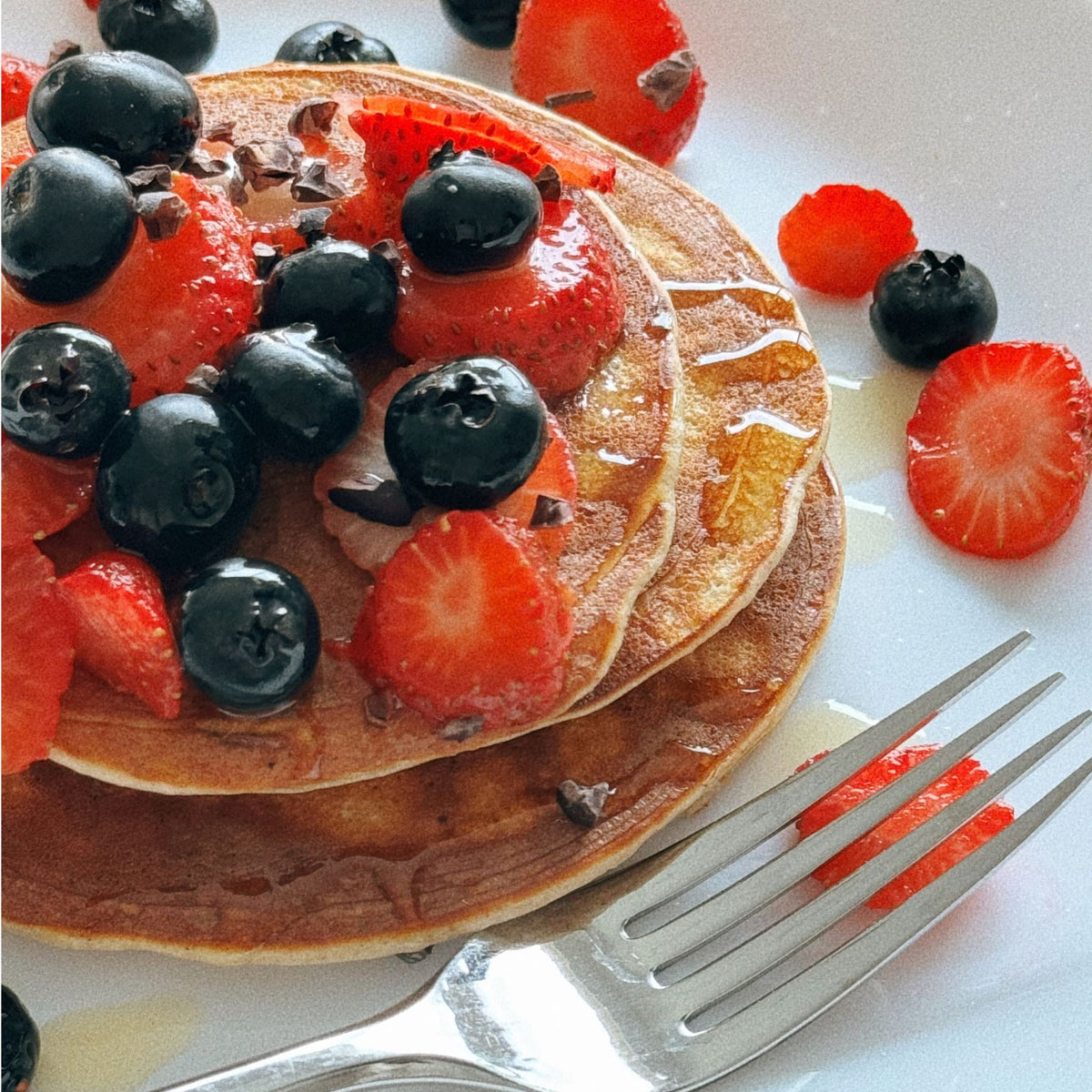 A close-up of a stack of golden pancakes topped with fresh blueberries, sliced strawberries, chocolate pieces, and a drizzle of syrup, served on a white plate with a fork.