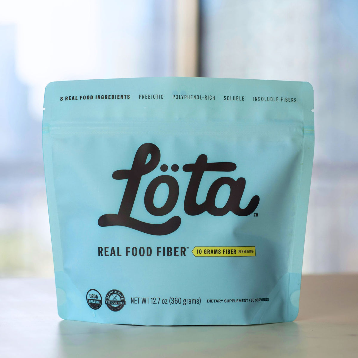 A light blue stand-up pouch of ORGANIC Löta Real Food Fiber dietary supplement, featuring the brand name 'Löta' in large black script, with 'Real Food Fiber' and '10 Grams Fiber Per Serving' printed below, set on a light surface with a blurred indoor background. Prebiotic Fiber, Polyphenol-Rich Fiber, Soluble Fiber, Insoluble Fiber. Chicago blurred skyline. Glyphosate Residue Free Fiber. Lota (Löta)