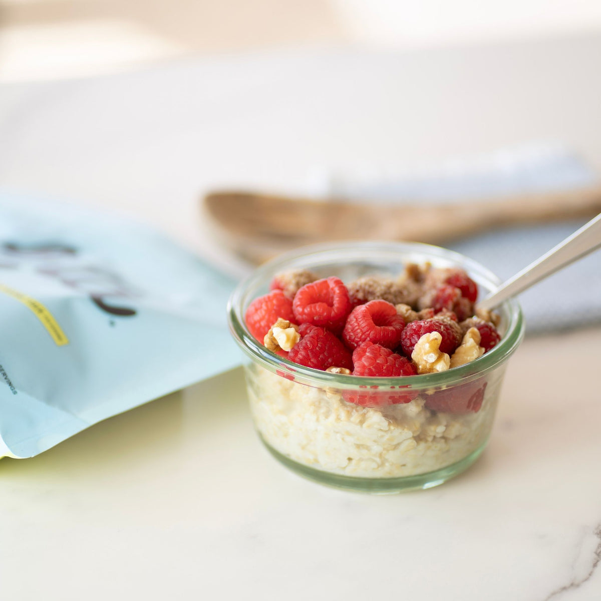 A small glass bowl filled with oatmeal topped with fresh raspberries and nuts, with a spoon resting inside. A light blue pouch is blurred in the background on a light marble surface. Löta real food fiber pouch. A light blue stand-up pouch of ORGANIC Löta Real Food Fiber dietary supplement, featuring the brand name 'Löta' in large black script, with 'Real Food Fiber' and '10 Grams Fiber Per Serving' printed below, set on a light surface with a blurred indoor background. Prebiotic Fiber, Polyphenol-Rich Fiber