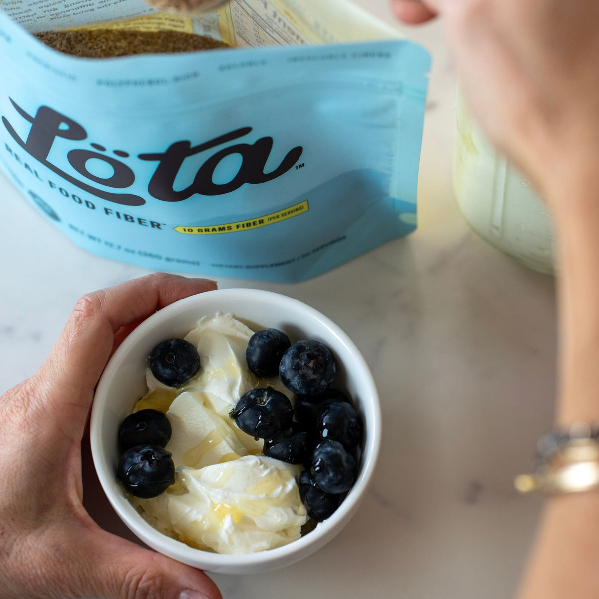 A person holds a small white bowl filled with Greek yogurt topped with blueberries, with a large blue bag of Löta organic real food fiber in the background on a light marble surface. Gold jewelry bracelets. A light blue stand-up pouch of ORGANIC Löta Real Food Fiber dietary supplement, featuring the brand name 'Löta' in large black script, with 'Real Food Fiber' and '10 Grams Fiber Per Serving' printed below, set on a light surface with a blurred indoor background. Prebiotic Fiber, Polyphenol-Rich Lota 